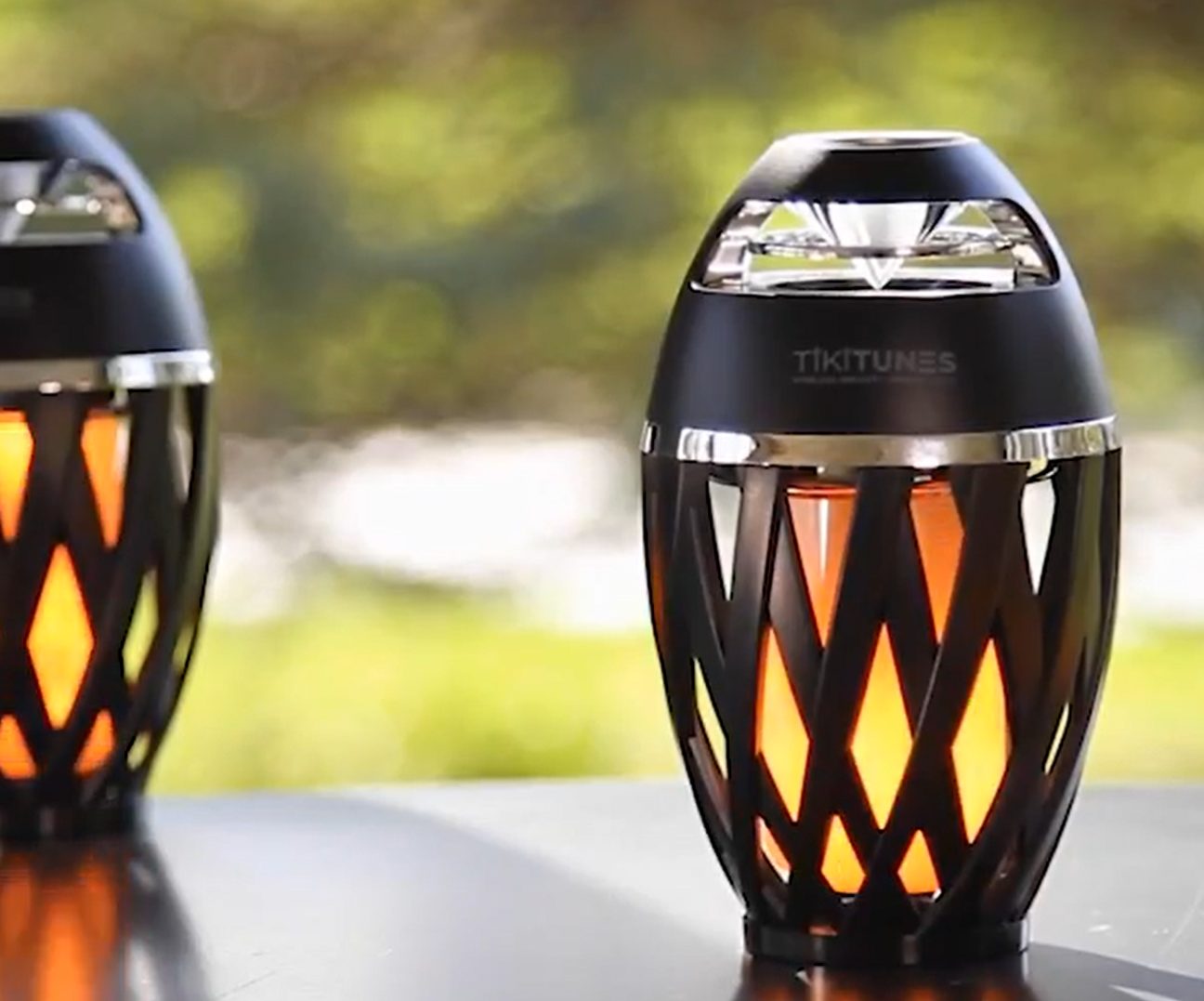Two TikiTunes wireless speakers with a warm flame-like glow, one in front of the other on a dark outdoor table